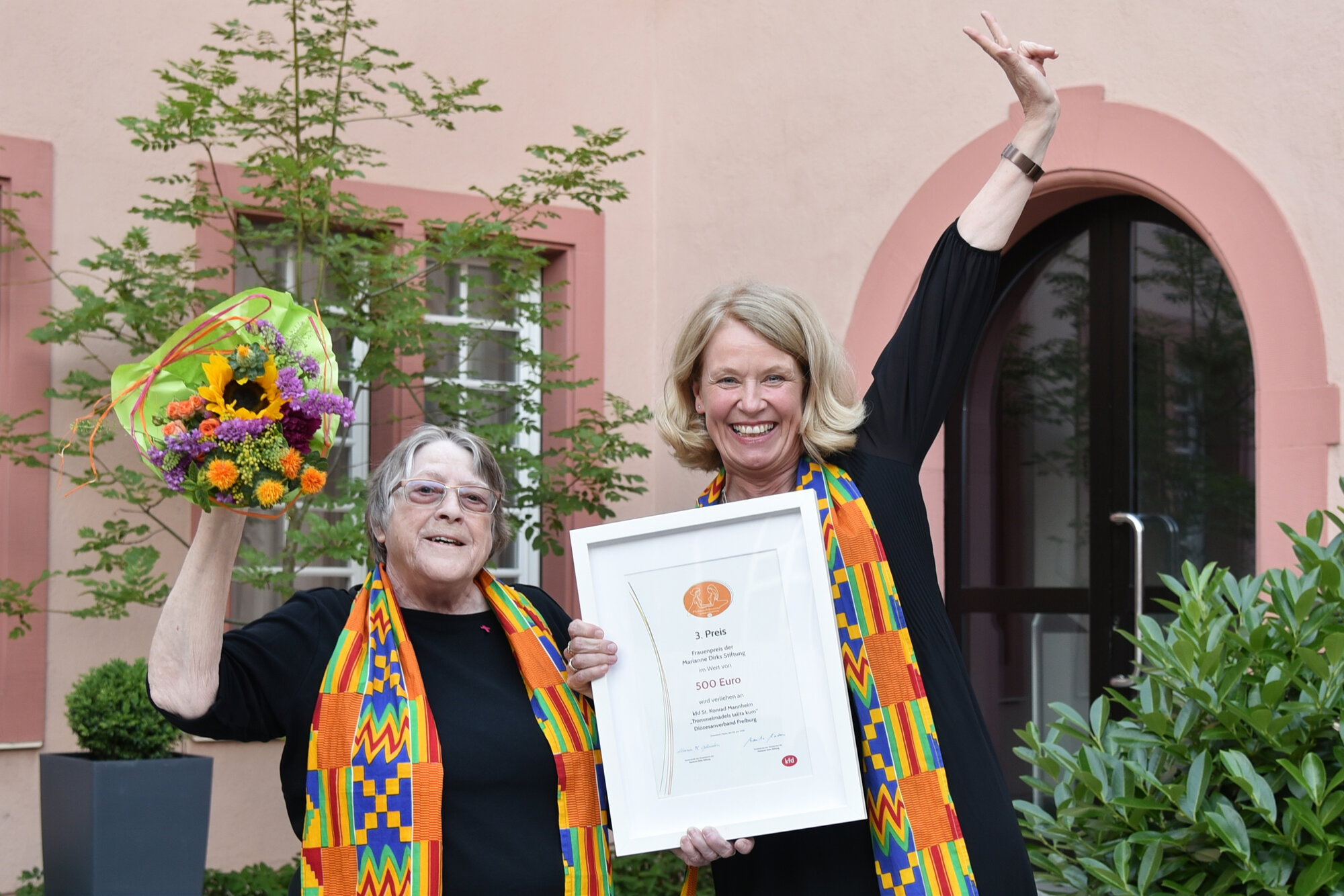 Zwei Frauen freuen sich und halten eine große Urkunde und einen Blumenstrauß hoch